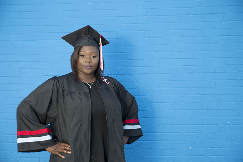 senior posing in front of blue painted brick wall in cap and gown